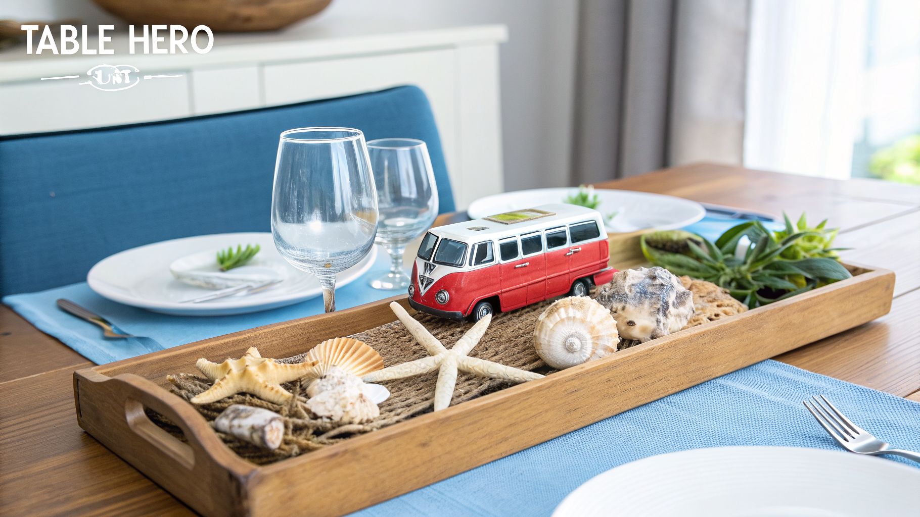 Coastal-themed dining table centerpiece with a red toy bus, starfish, seashells, and greenery on a wooden tray.
