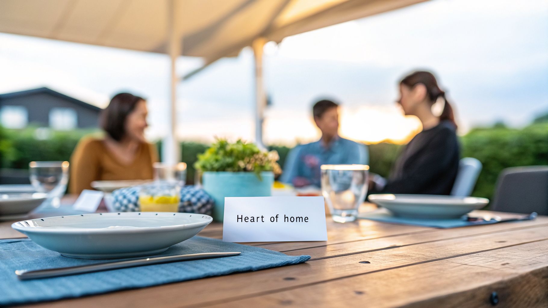 A cozy outdoor dining table setting with tableware, a plant centerpiece, and 'Heart of home' sign, with blurred guests.