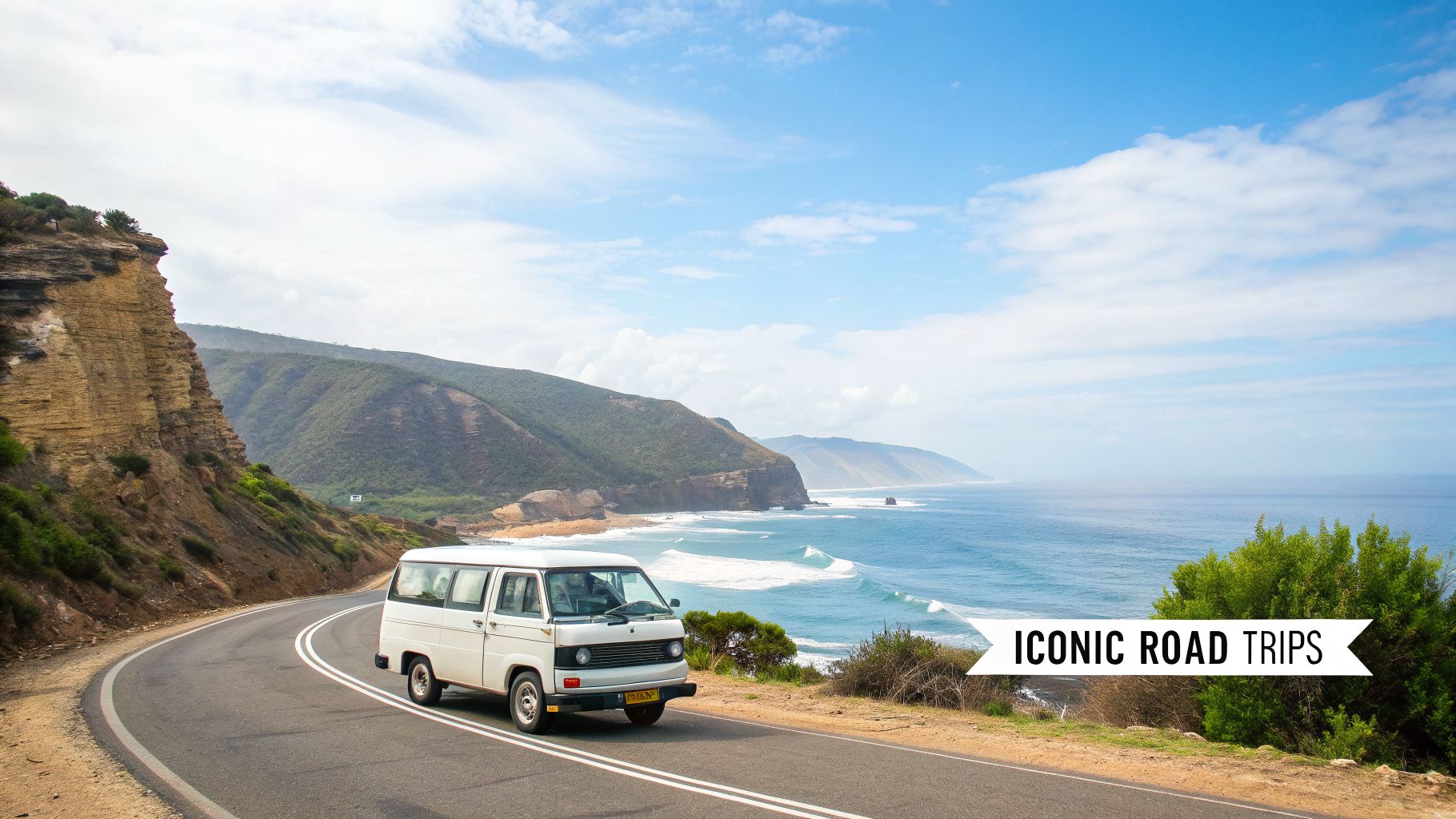 A white camper van drives along a stunning coastal road beside the blue ocean and green hills.