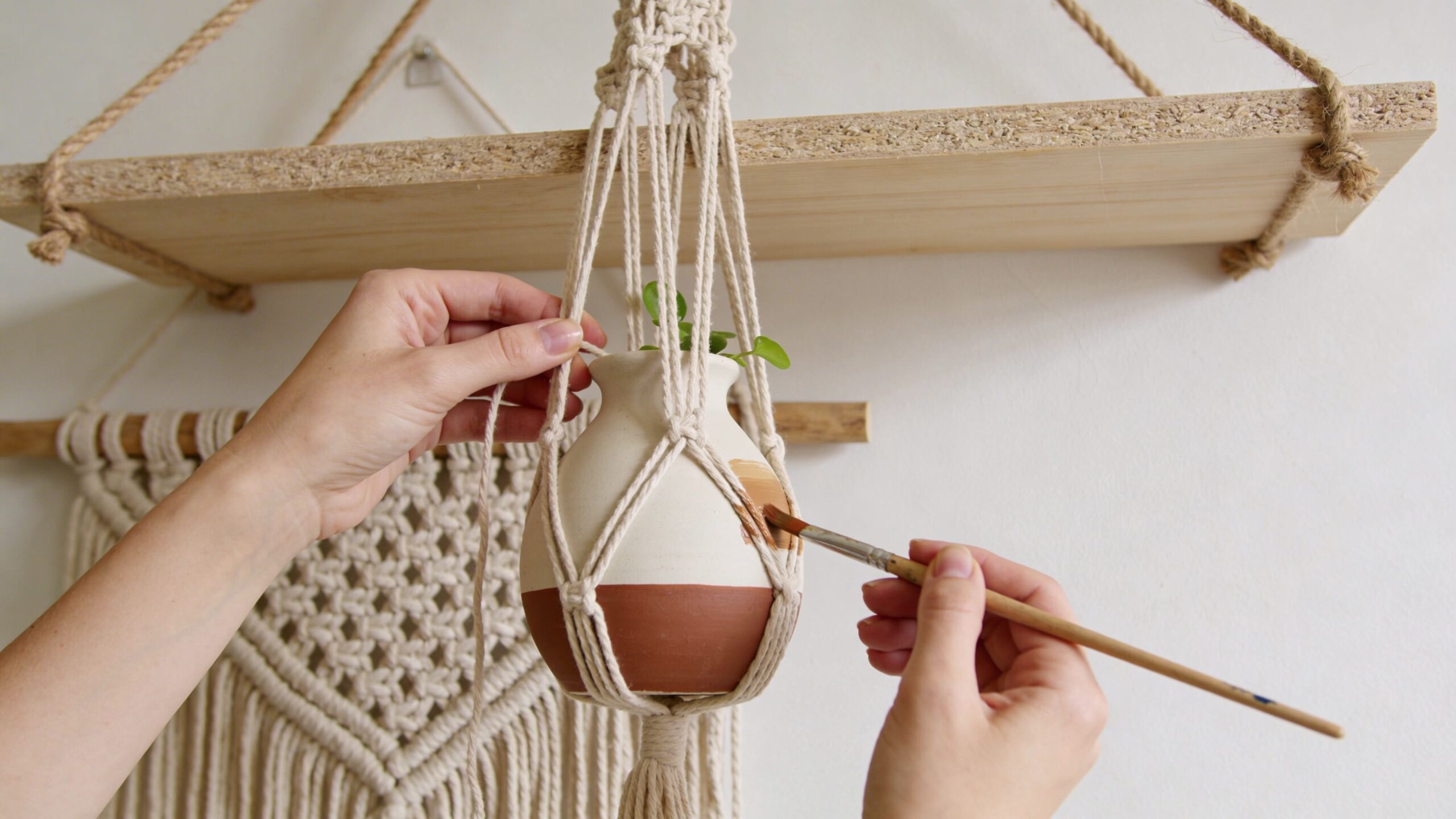 A person painting a small terracotta planter while it hangs in a decorative macrame plant hanger.