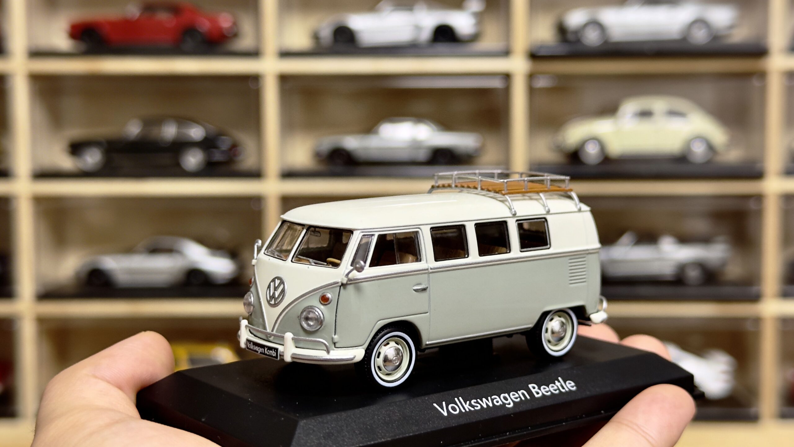 A hand holding a model of a vintage two-tone Volkswagen bus against a background of toy cars.