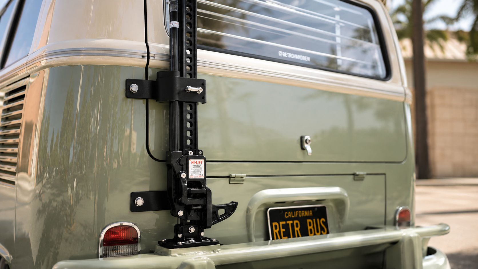 A black Hi-Lift jack mounted to the rear exterior of a vintage sage green Volkswagen bus.