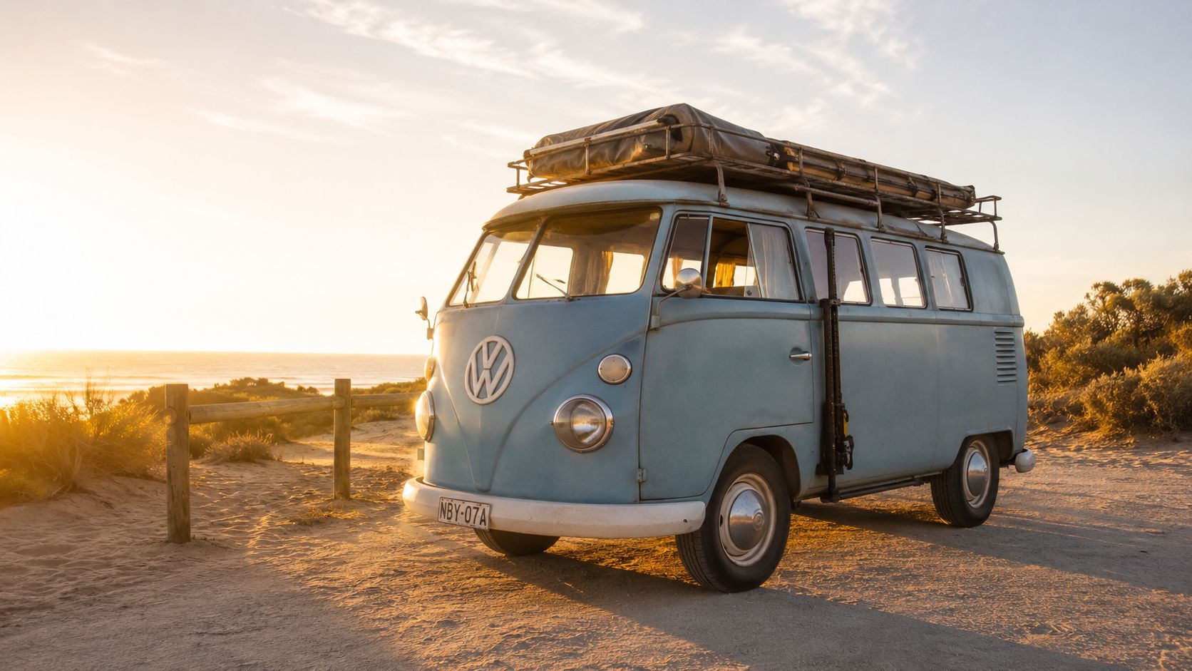 A vintage light blue Volkswagen bus parked on a sandy beach during a beautiful golden sunset.