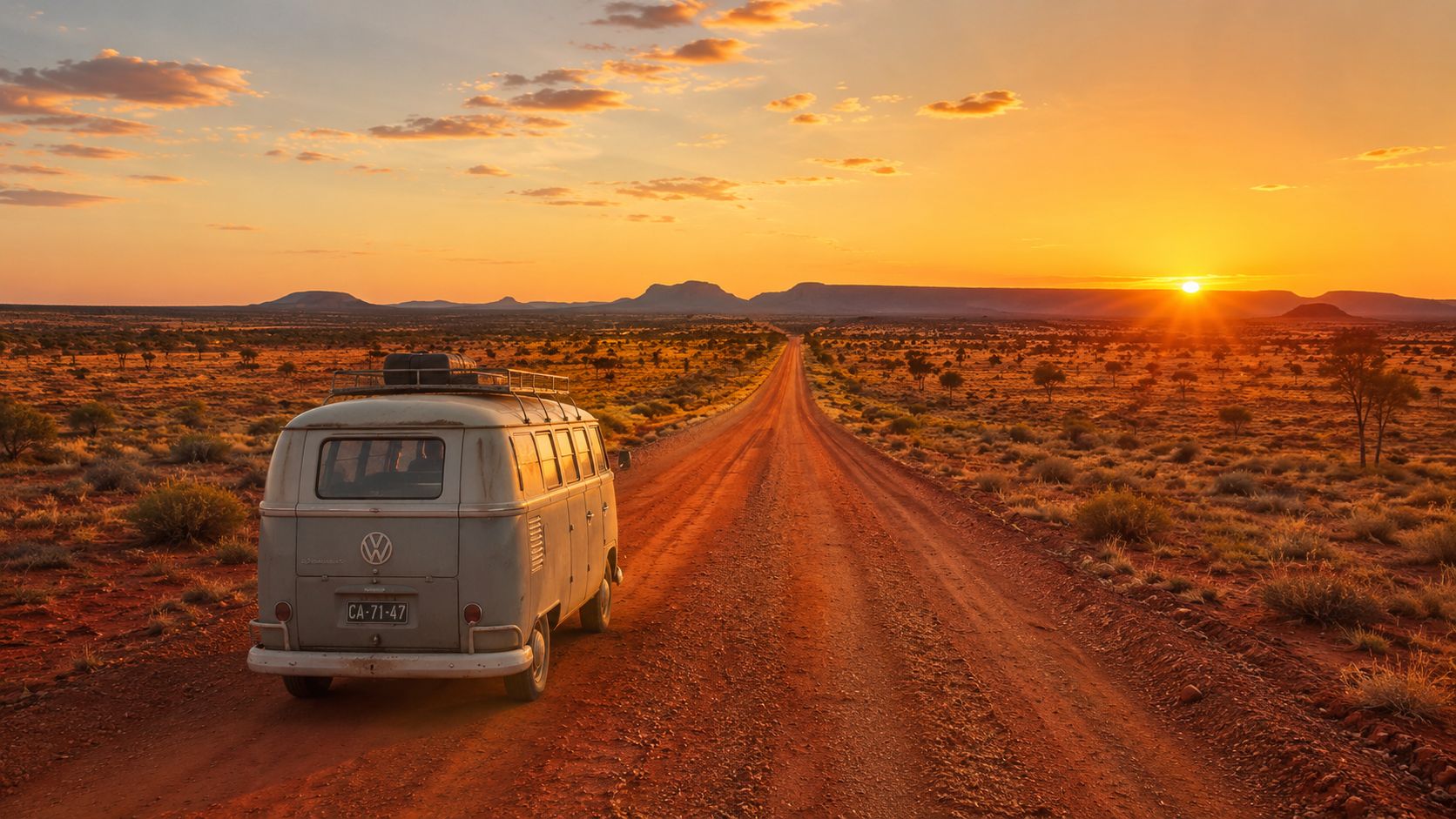A vintage Volkswagen bus driving on a dusty red road through the vast Australian outback at sunset.