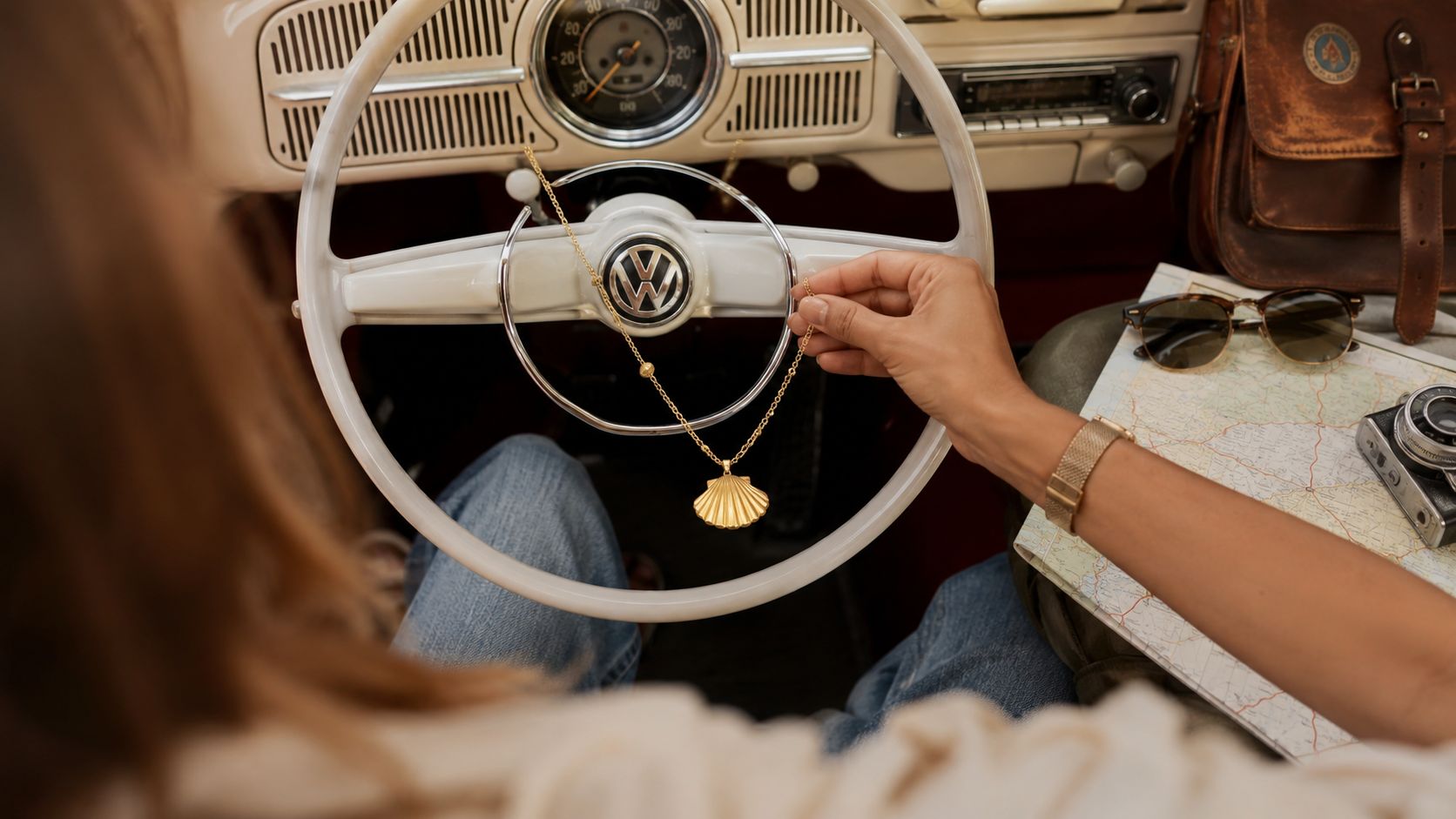 A person holds a delicate gold shell necklace inside a vintage Volkswagen car with a map nearby.