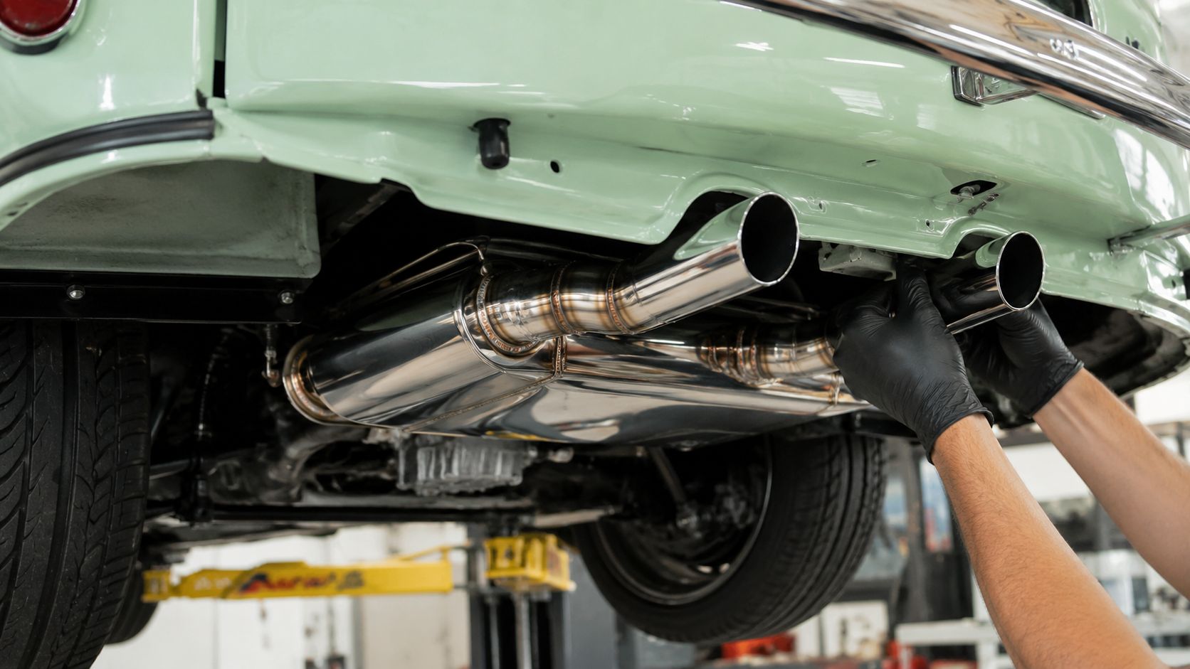 A mechanic wearing black protective gloves installs a shiny stainless steel exhaust system under a vintage green car.