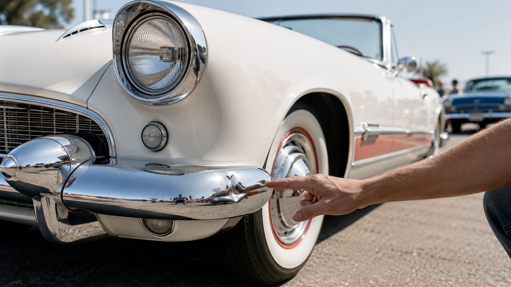A person pointing to a small dent on the shiny chrome bumper of a vintage white car.