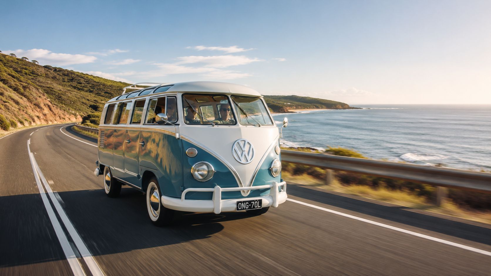 A vintage blue and white Volkswagen bus driving along a scenic coastal road during a bright day.