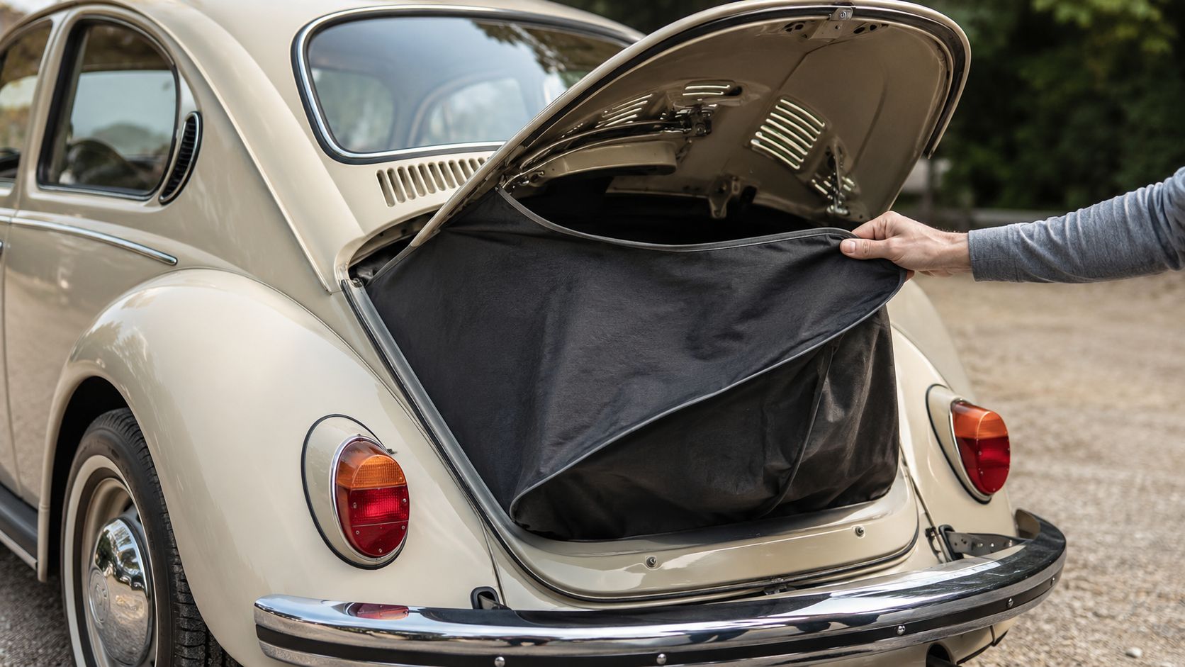 A person placing a black organizer bag into the trunk of a vintage beige Volkswagen Beetle car.