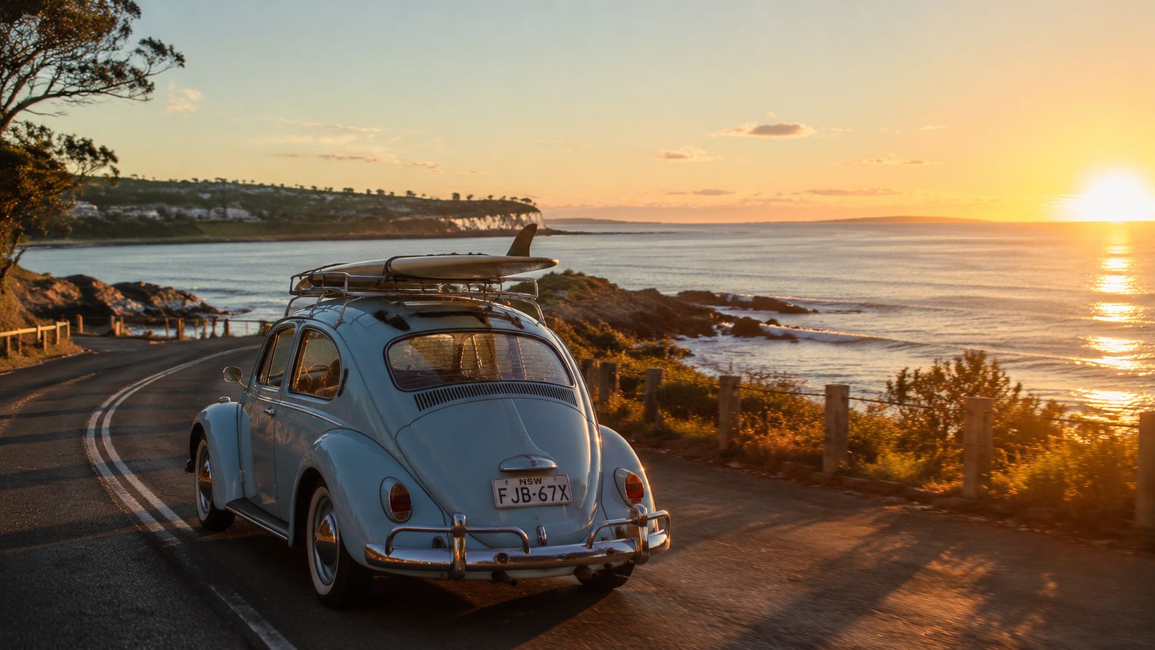 A vintage light blue Volkswagen Beetle with a surfboard on its roof driving along a coastal road.