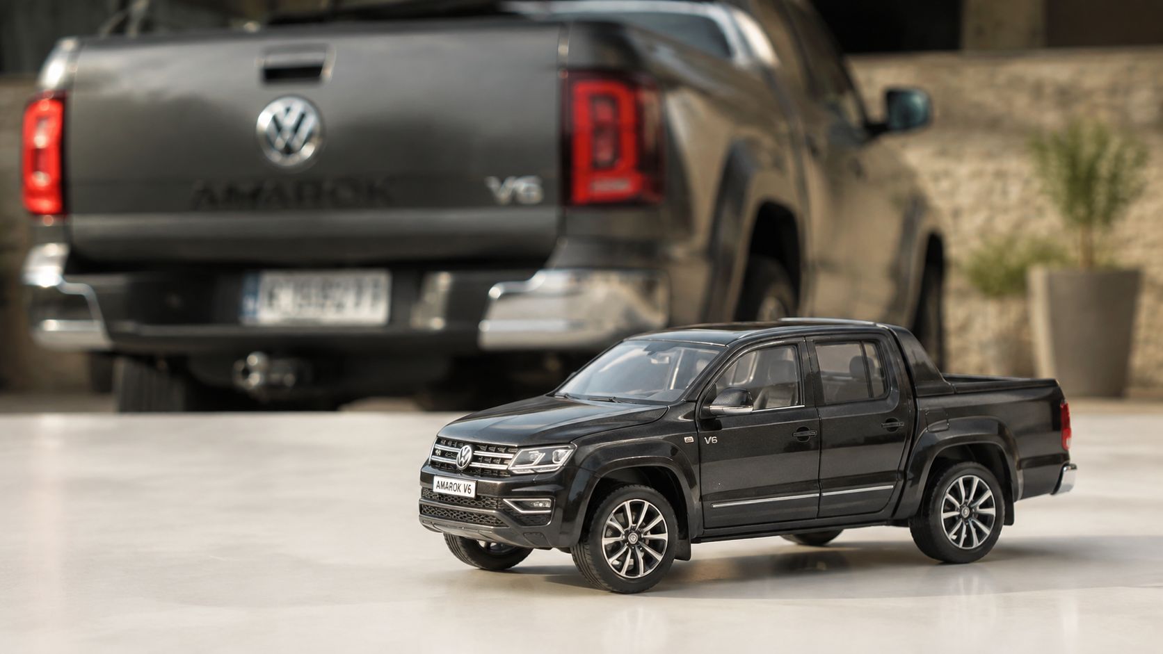 A miniature black Volkswagen Amarok V6 toy truck parked in front of a real full-sized Amarok vehicle.