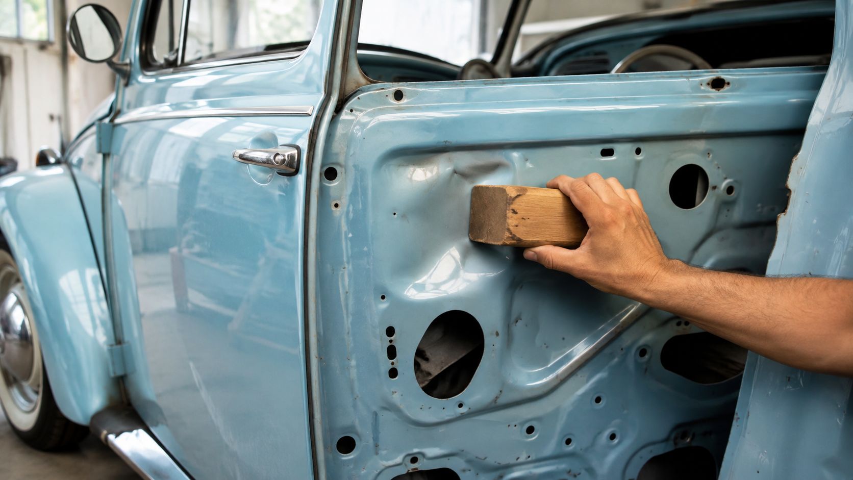 A car technician uses a wooden block to repair a dent on a light blue vintage car door.