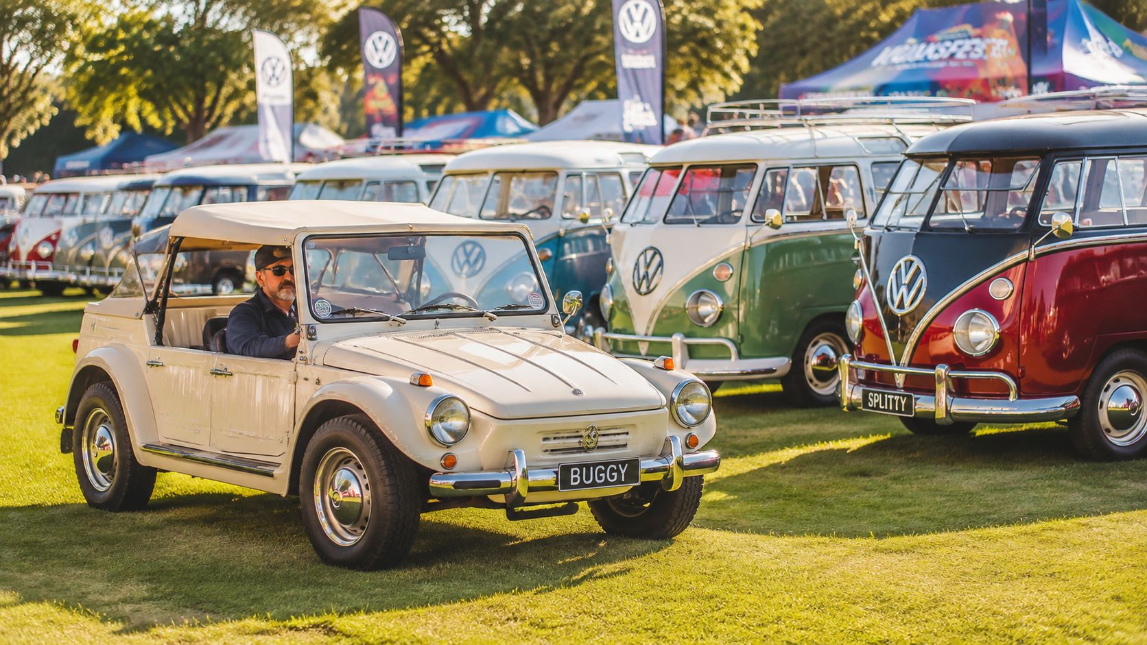 A classic beige Volkswagen buggy driving on a grassy field at a sunny car show event.