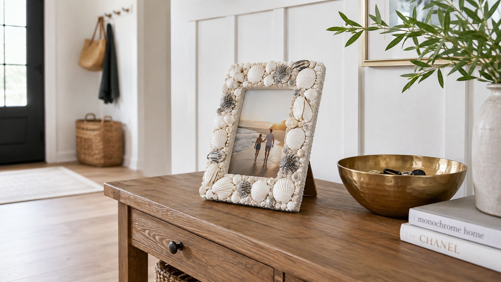 A coastal style seashell photo frame resting on a wooden console table with a brass bowl nearby.