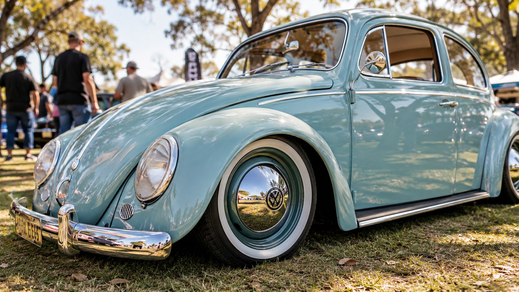 A vintage light blue Volkswagen Beetle parked on grass with prominent chrome hubcaps and whitewall tires.
