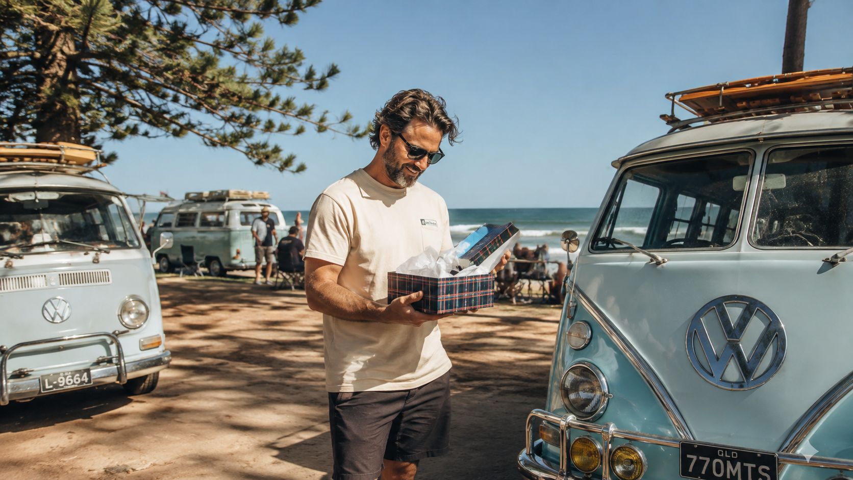 A bearded man smiling while opening a gift box between two vintage Volkswagen campervans at the beach.
