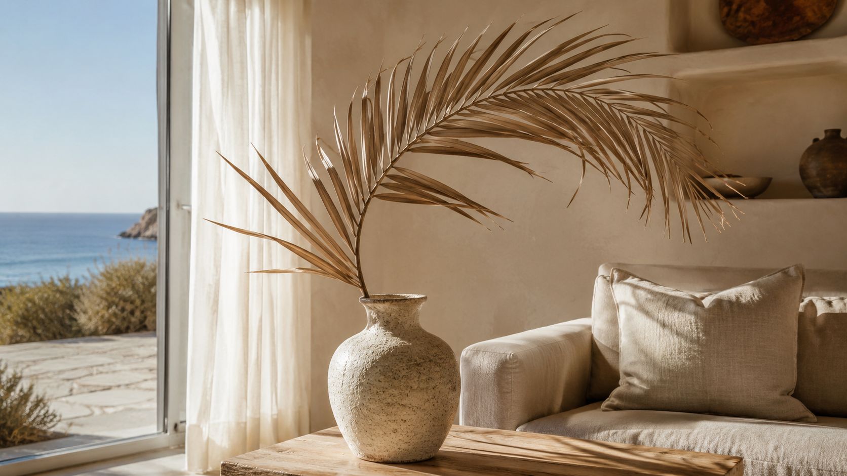 A dried palm leaf in a textured ceramic vase sits on a wooden table beside a sofa.